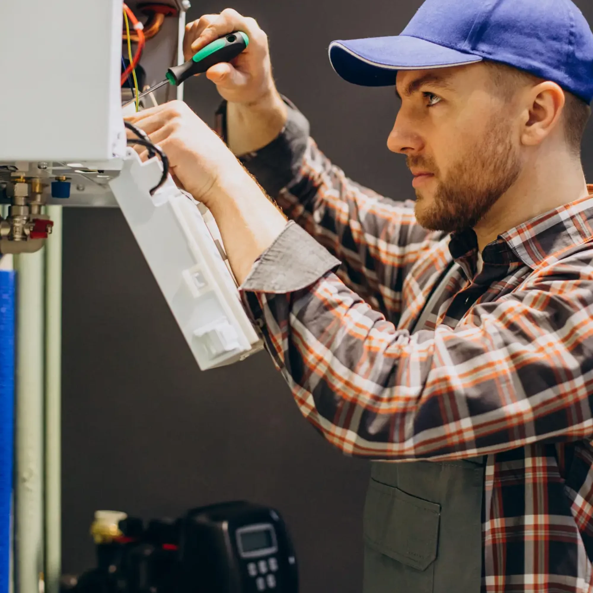 chauffagiste en casquette bleue effectuant un branchement électrique à l’intérieur d’une chaudière murale ouverte — maintenance et vérification des connexions du système de chauffage.